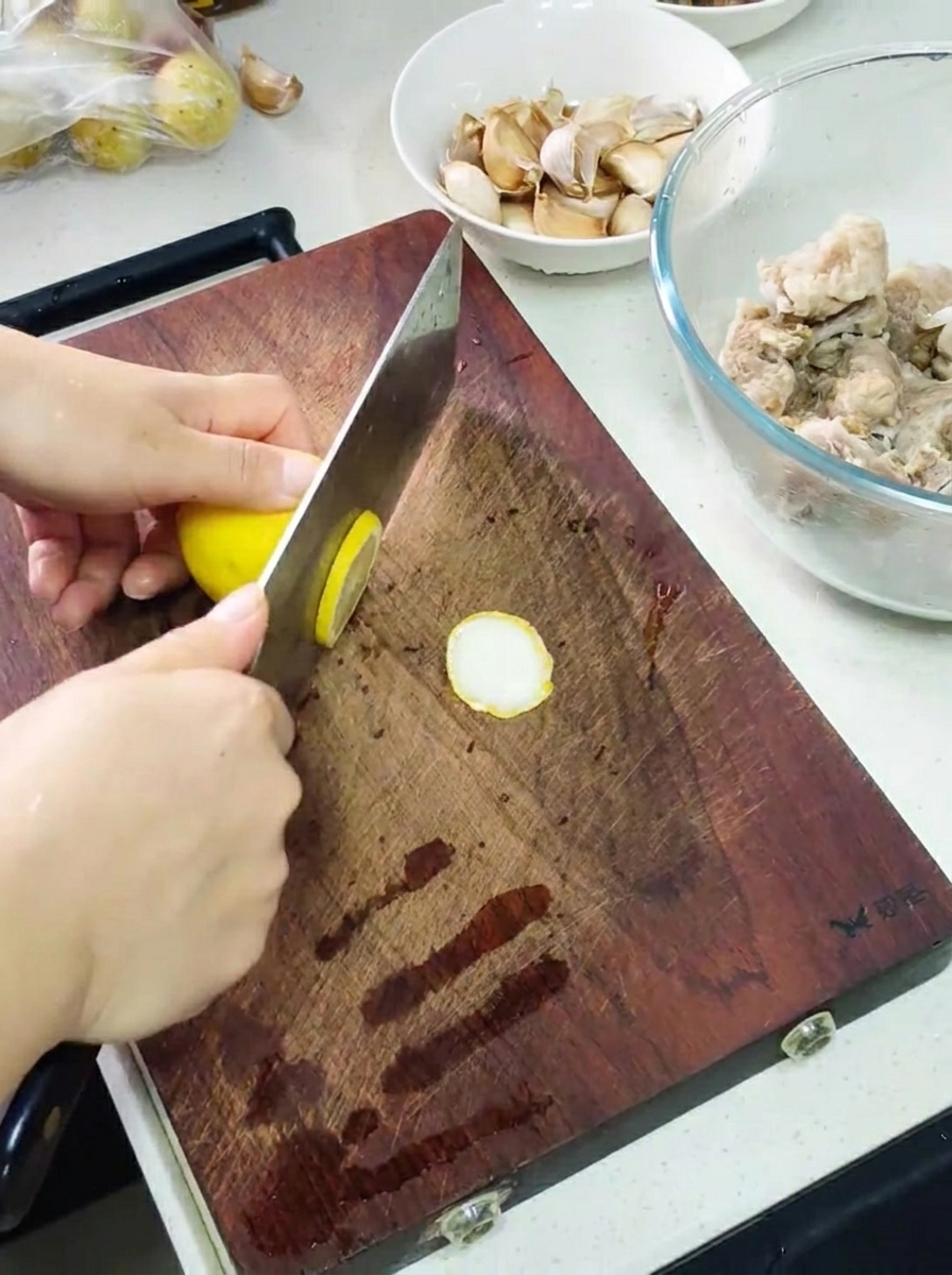 Mani che usano una mannaia per affettare un limone su un tagliere, con una ciotola di costine marinate e spicchi daglio nelle vicinanze.