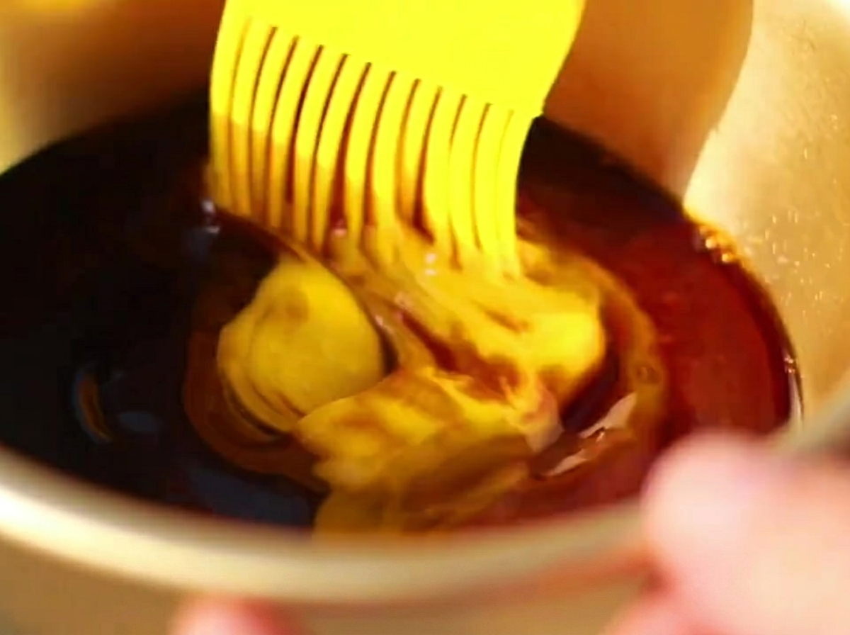 Un pennello in silicone che applica una salsa scura e ricca sulla superficie delle costine di maiale parzialmente cotte in una teglia.