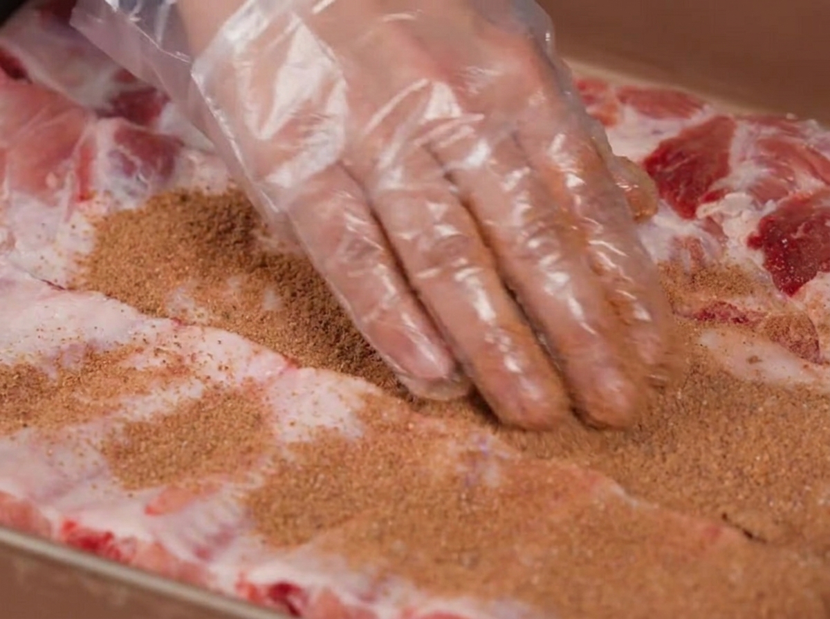 Mani che indossano guanti di plastica massaggiano una miscela di spezie marroni su costine di maiale crude.
