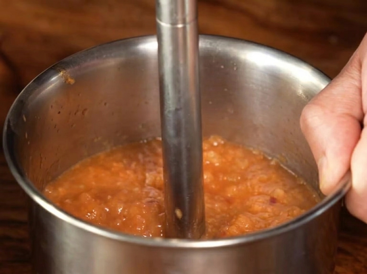 Un frullatore a immersione in metallo che lavora la salsa cotta riducendola in una purea liscia.