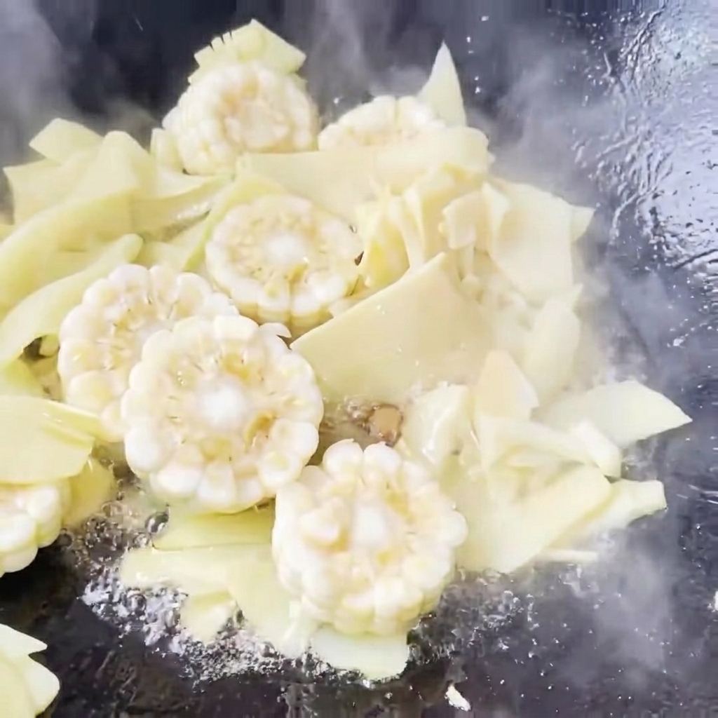 Acqua limpida versata in un wok che contiene mais e germogli di bambù saltati, iniziando a sobbollire.