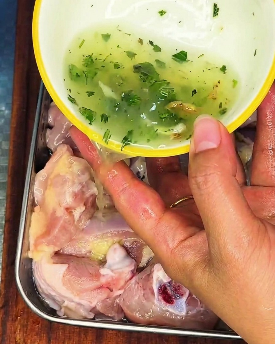 Una mano versa un liquido con cipollotto e zenzero sul pollo crudo in un contenitore di acciaio inossidabile.