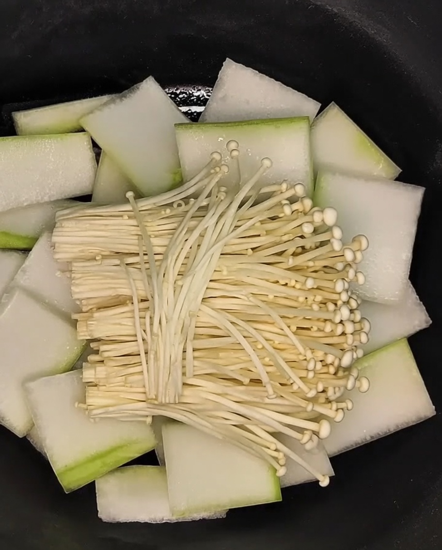 Un mazzetto di funghi enoki appoggiato sul letto di fette di melone invernale nella pentola.