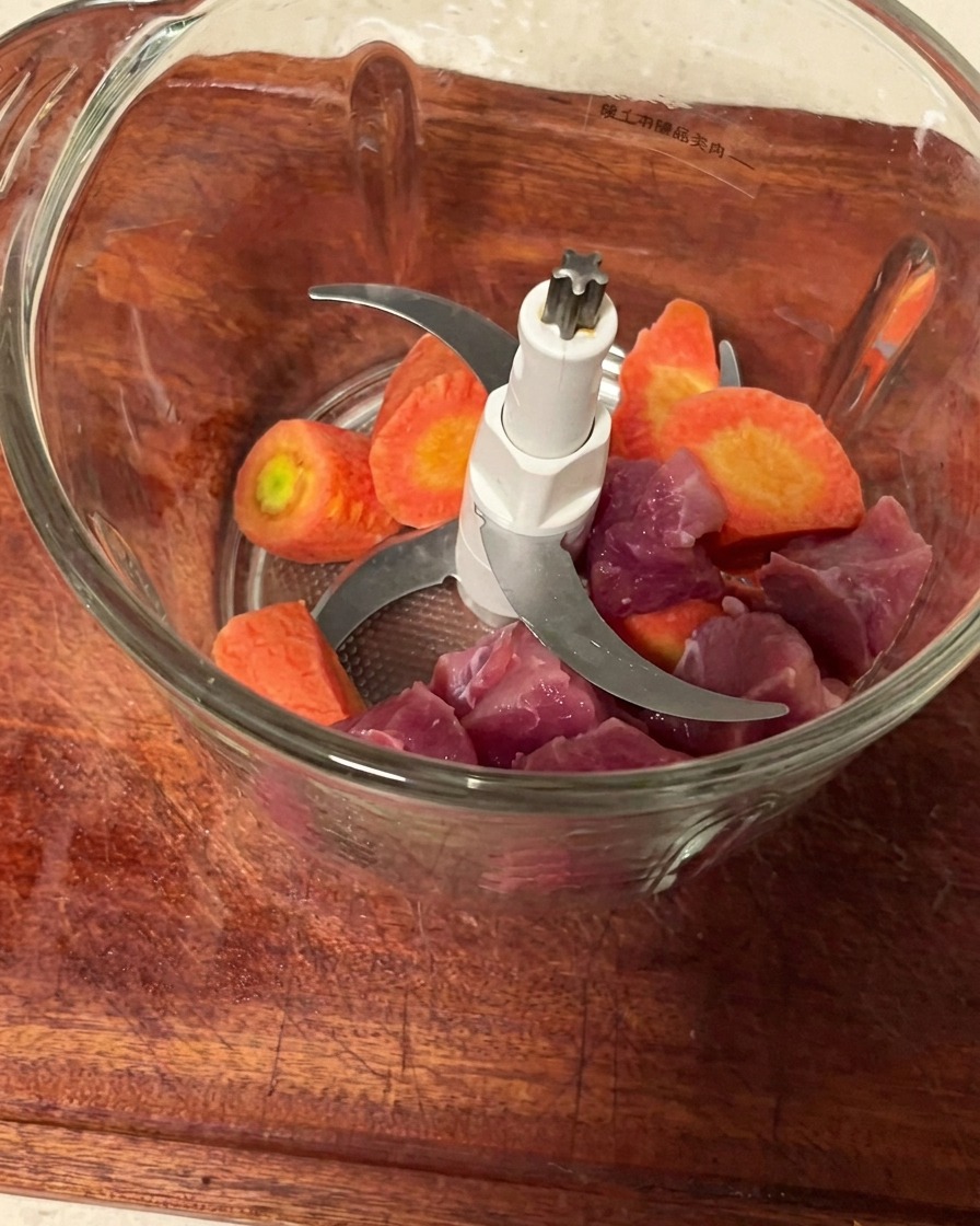 Pezzi di maiale e cubetti di carota allinterno di un contenitore di un tritacarne pulito, pronti per essere macinati.