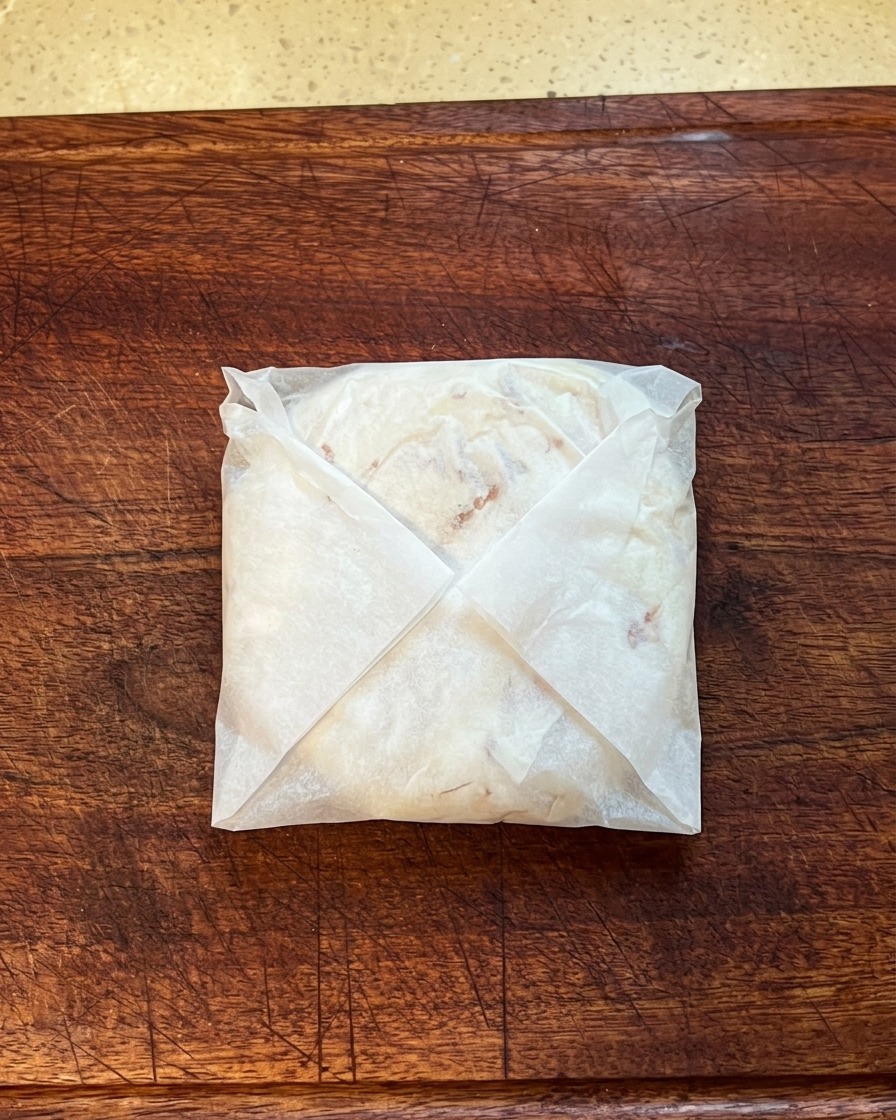 Un pancake allo scalogno ripiegato e racchiuso in carta da forno, mentre viene schiacciato su un tagliere di legno.