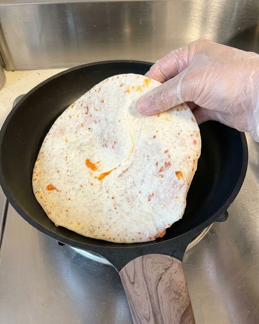 Una mano che indossa un guanto di plastica posiziona un pancake appiattito e avvolto nella carta da forno in una padella antiaderente nera calda.