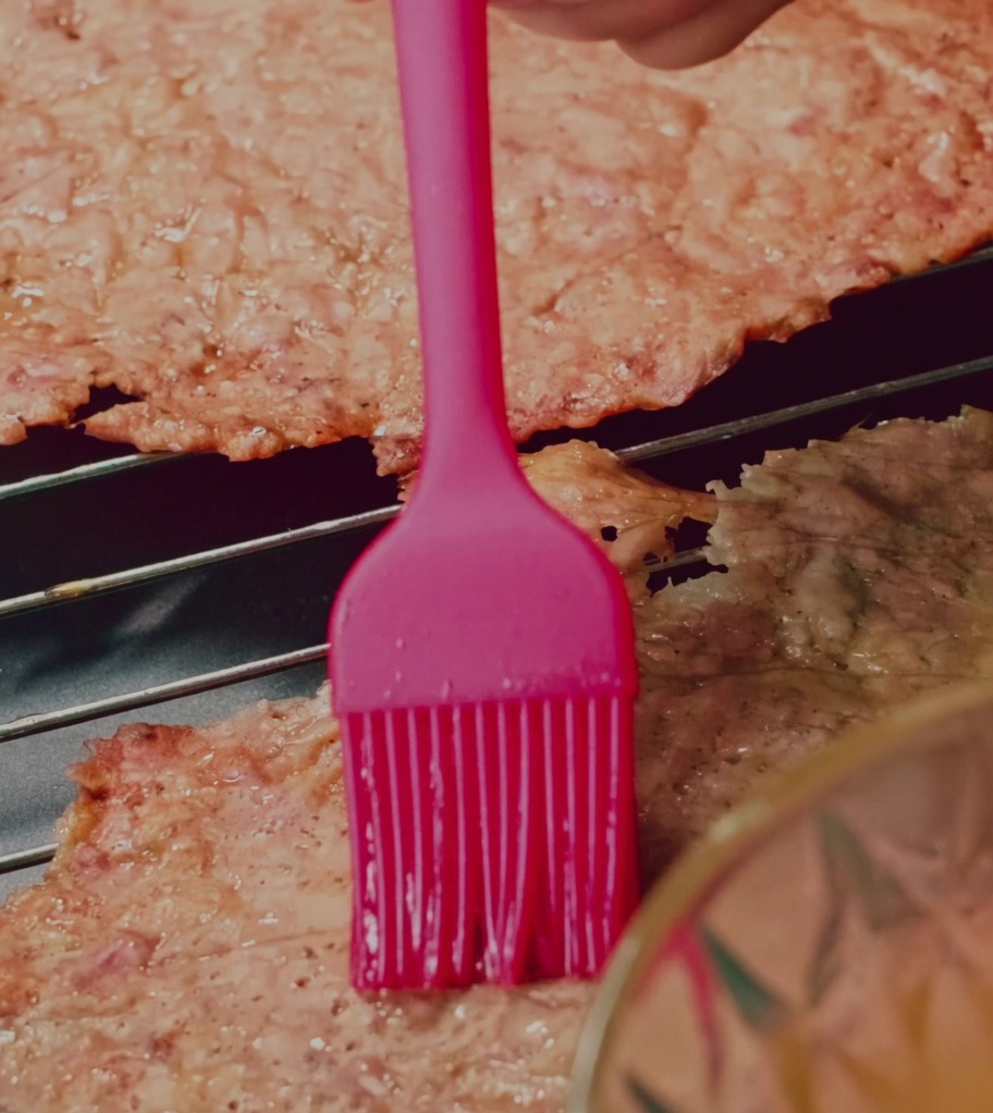 Un pennello in silicone applica una glassa di miele e acqua sulle fette di jerky di maiale su una griglia.