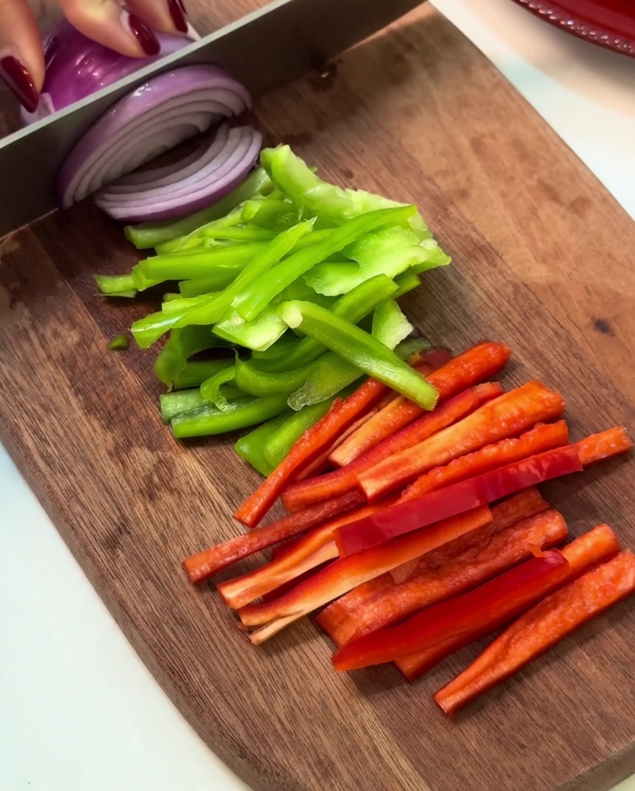 Strisce di peperone verde, peperone rosso e cipolla viola disposte ordinatamente su un tagliere di legno.