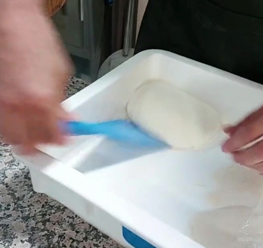 Una porzione di impasto per pizza sollevata da un contenitore con una spatola in plastica blu.