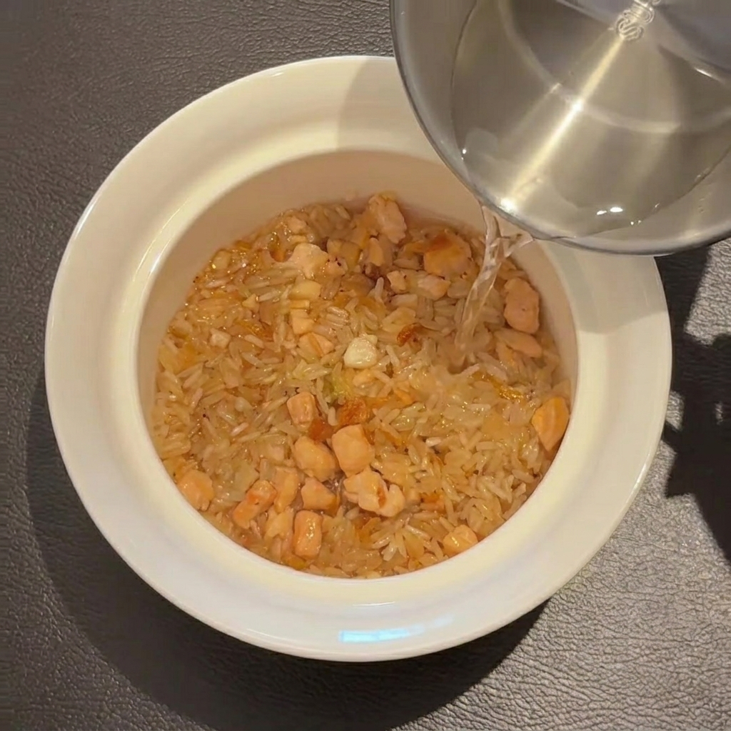 Acqua versata da una tazza in acciaio inossidabile sul composto di riso in una ciotola bianca.