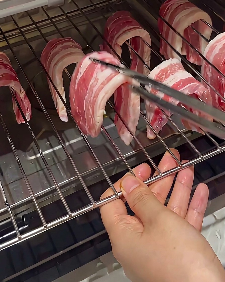 Fette di pancetta cruda appese ai fili di una griglia da forno, tenute con le pinze.