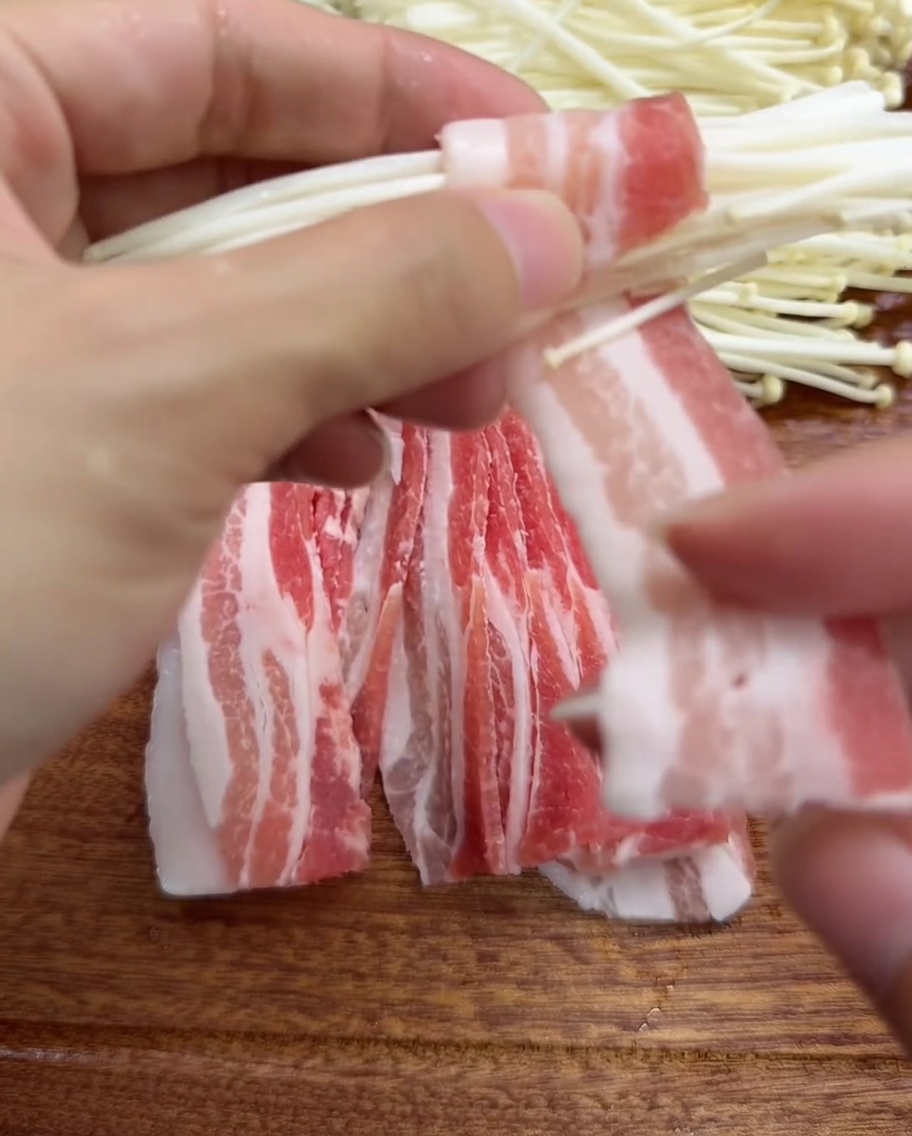 Mani che avvolgono una fetta sottile di pancetta attorno a un mazzetto di funghi enoki.