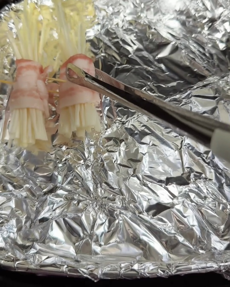 Involtini di pancetta ed enoki posizionati nel cestello della friggitrice ad aria rivestito di carta stagnola.