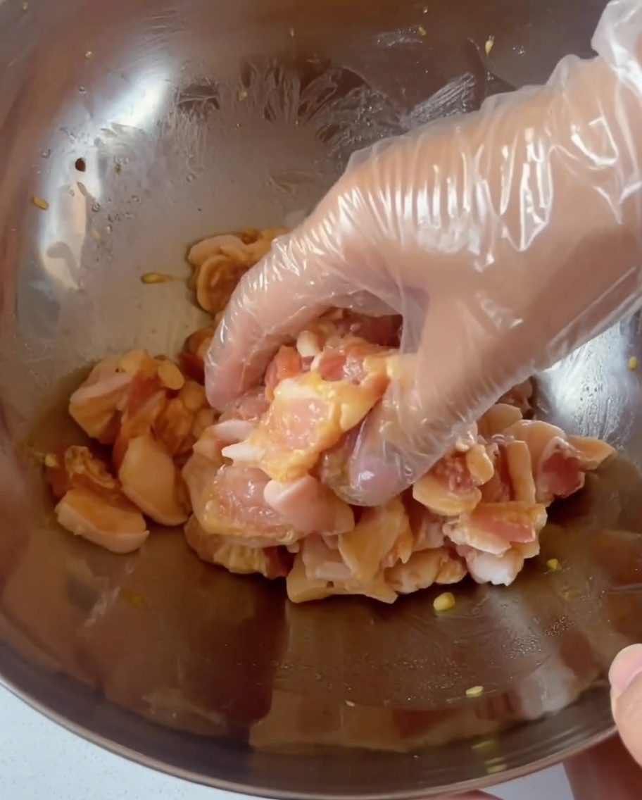 Una mano con un guanto che mescola le fette di maiale condite in una grande ciotola in acciaio inossidabile.