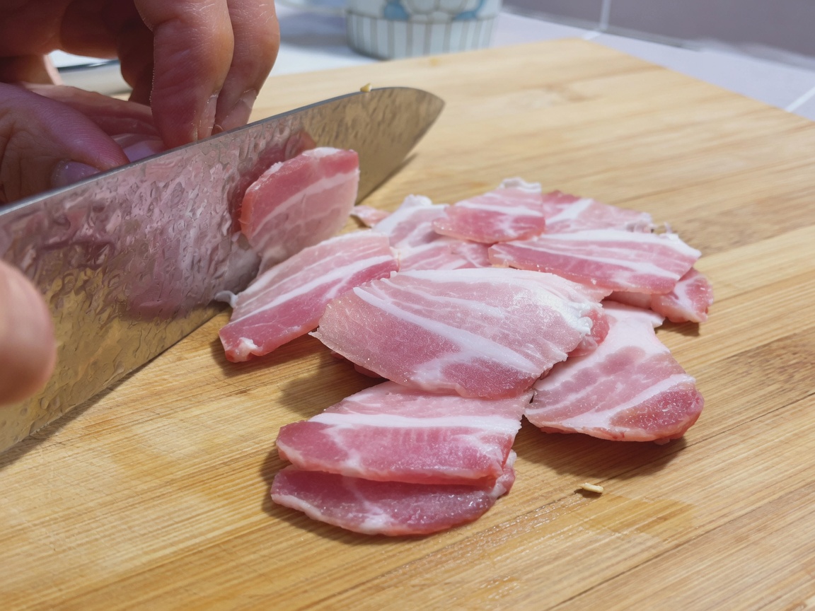 Sottili fette di pancetta cruda disposte su un tagliere di legno accanto a un grande coltello da chef.