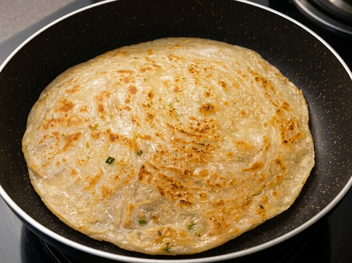 Un pane piatto rotondo allo scalogno che frigge in una padella antiaderente scura, mostrando una superficie dorata e strutturata.