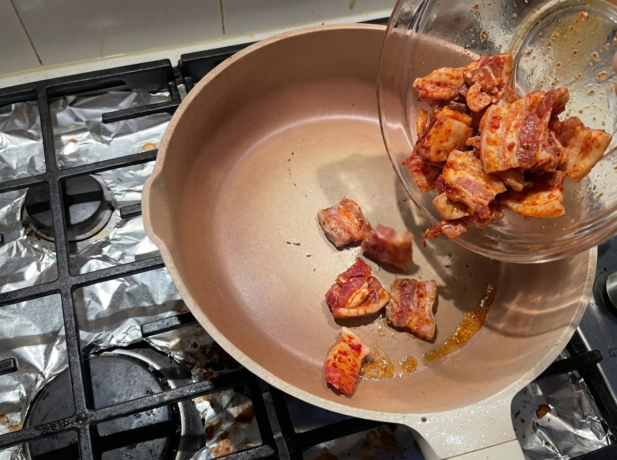Fette di pancetta marinata che scivolano da una ciotola di vetro in una padella calda e vuota per far sciogliere il grasso.