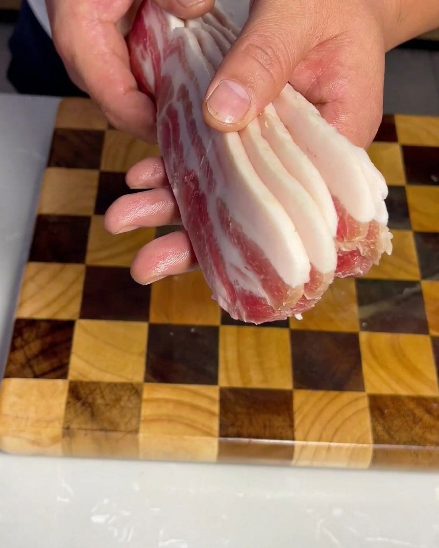 Strisce uniformi di pancetta cruda disposte ordinatamente su un tagliere di legno.
