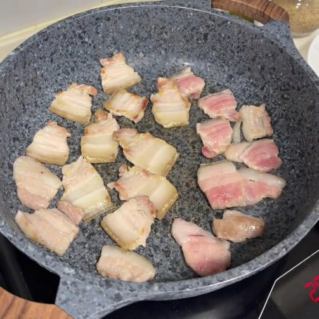 Fette di pancetta che friggono e dorano in una padella di pietra grigia.
