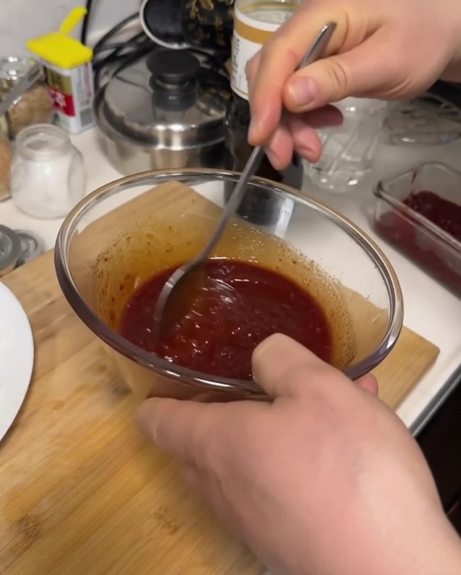 Una mano che usa un cucchiaio di metallo per mescolare una salsa densa e rosso scuro dentro una ciotola di vetro trasparente.
