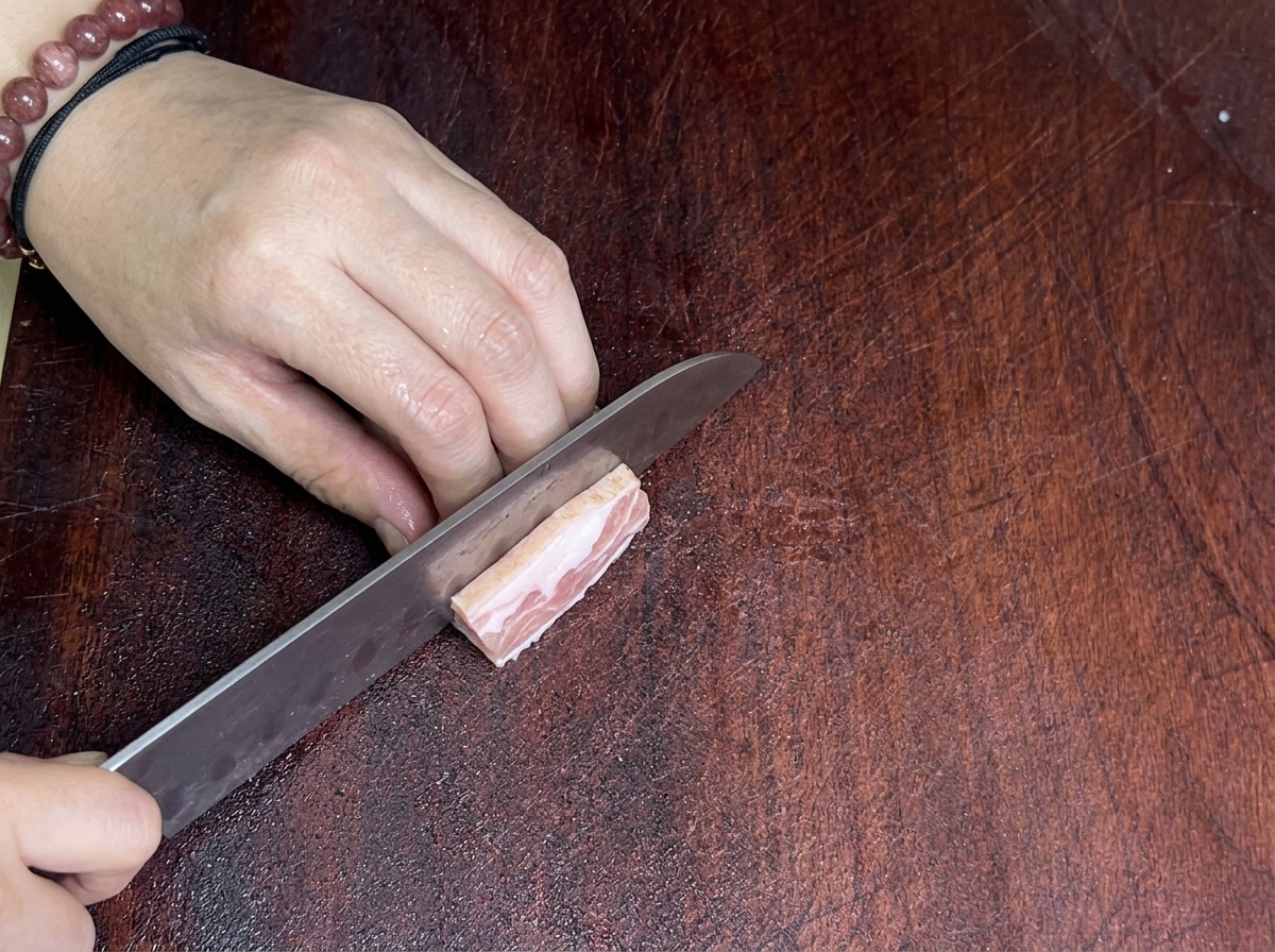 Un primo piano di un coltello affilato che taglia una fetta di pancetta su un tagliere di legno.