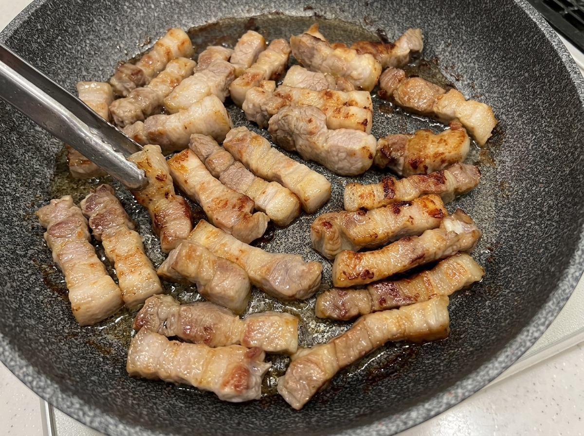 Pezzi di pancetta girati con le pinze in padella, mostrando superfici dorate.