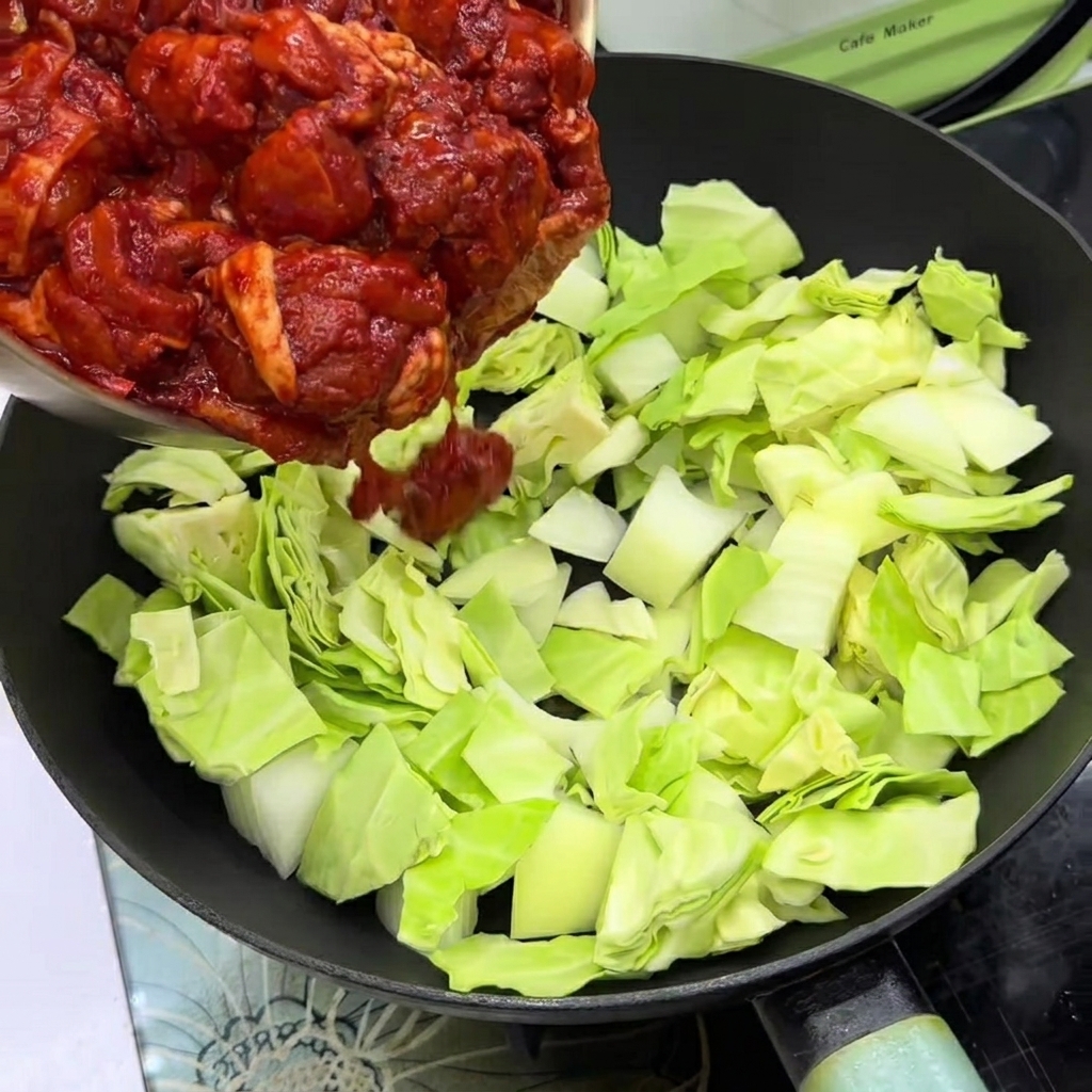 Pezzi di pollo marinati adagiati sopra cavolo e cipolle in una padella.
