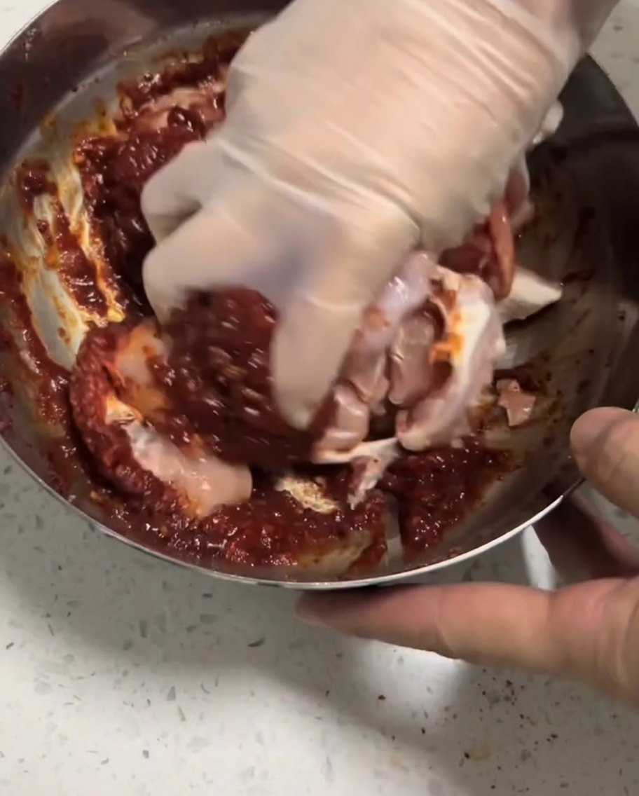 Pezzi di pollo crudo massaggiati con la marinatura rossa allinterno di una ciotola usando un guanto.