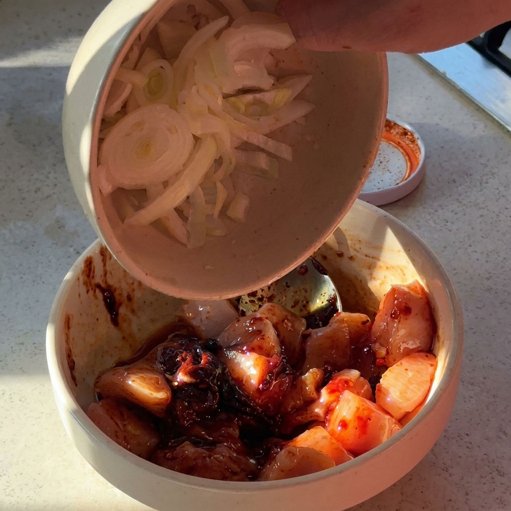 Cipolle affettate e aglio tritato aggiunti a una ciotola con i pezzi di pollo conditi.