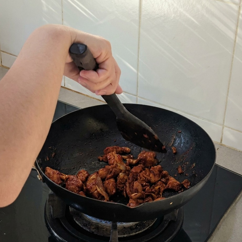 Pezzi di pollo e cipolla saltati in un wok nero con una spatola, ricoperti da una salsa scura e piccante.