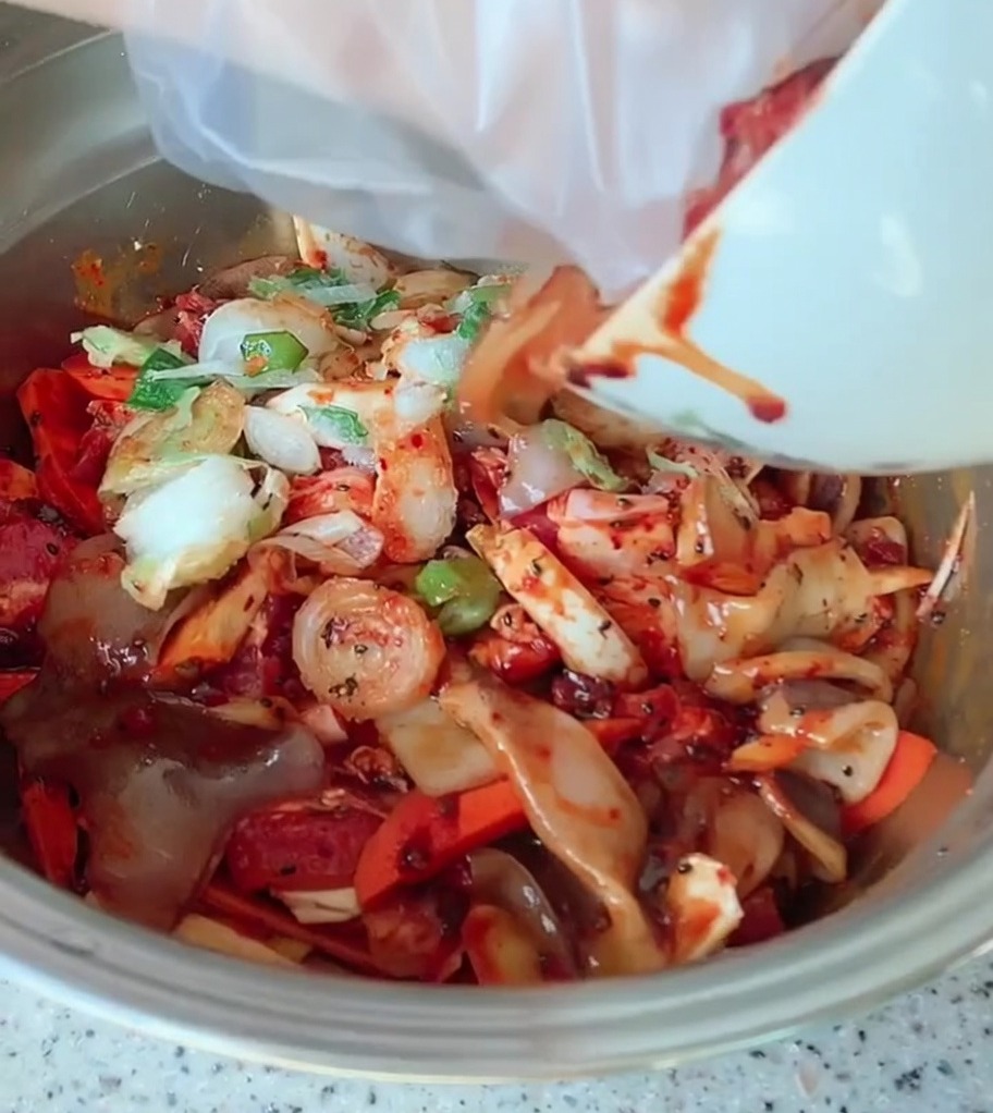 Una grande ciotola di metallo contenente fette di pancetta di maiale, verdure colorate e cipollotti ricoperti da una vivace salsa piccante rossa.