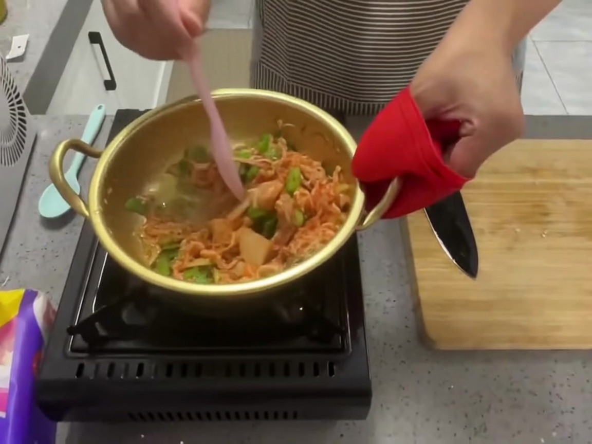 Kimchi, cipollotti e aglio mescolati insieme in una pentola con una spatola in silicone rosa.
