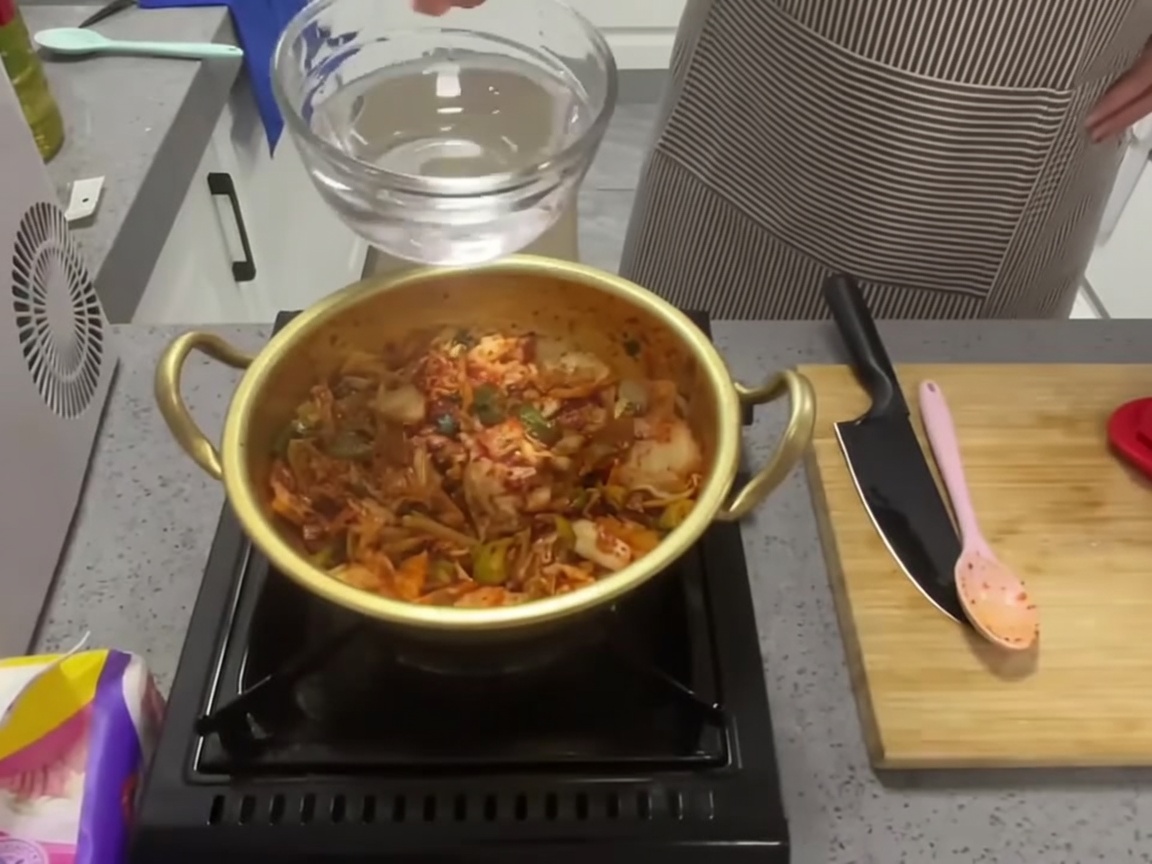 Acqua limpida versata da un misurino in una pentola dorata contenente kimchi saltato e aromi.