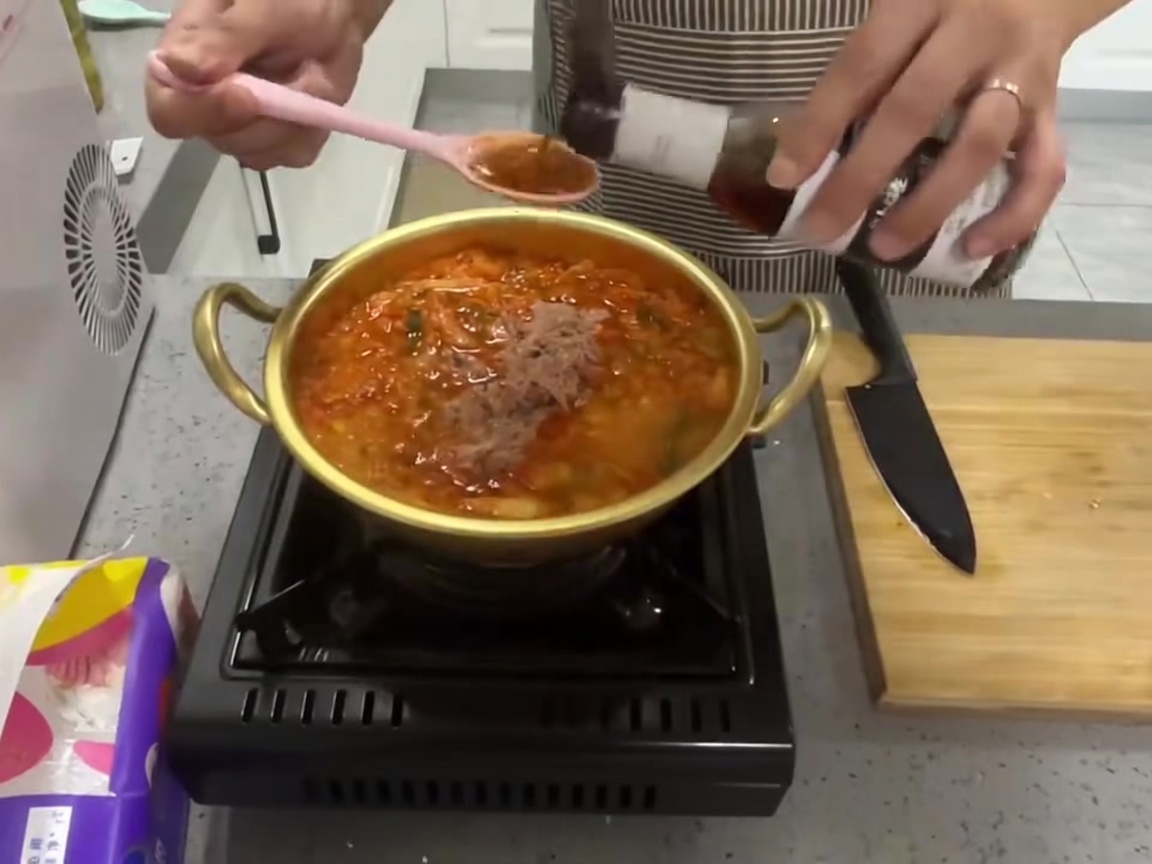 Un cucchiaio di condimento liquido scuro versato in una pentola di stufato bollente di kimchi e tonno.