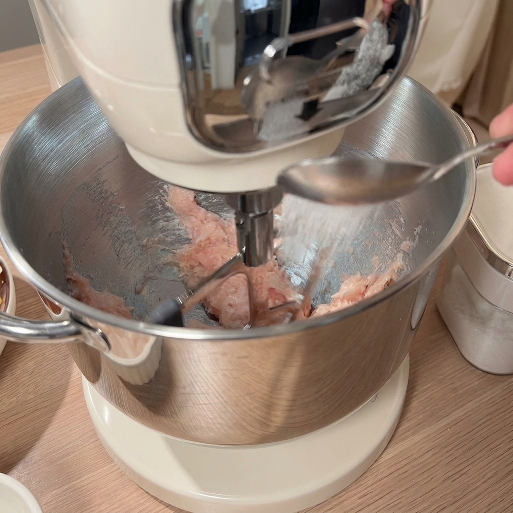 Un cucchiaio di condimento aggiunto al pollo macinato nella ciotola dellimpastatrice, con altri ingredienti visibili sul tavolo.