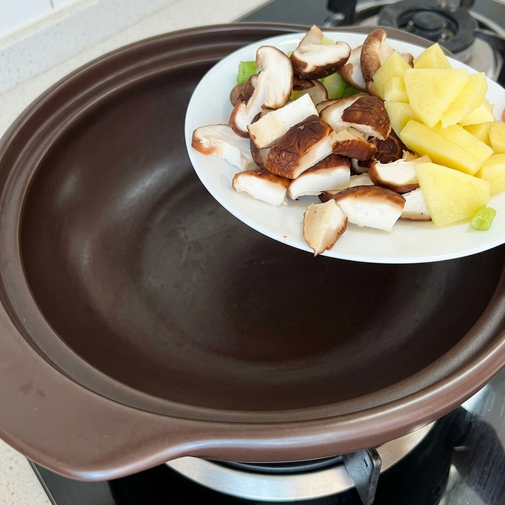 Cubetti di patate, zucchine e shiitake aggiunti in un tegame di terracotta.