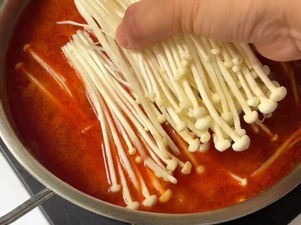 Un mazzetto di funghi enoki freschi aggiunto al brodo di kimchi in ebollizione.