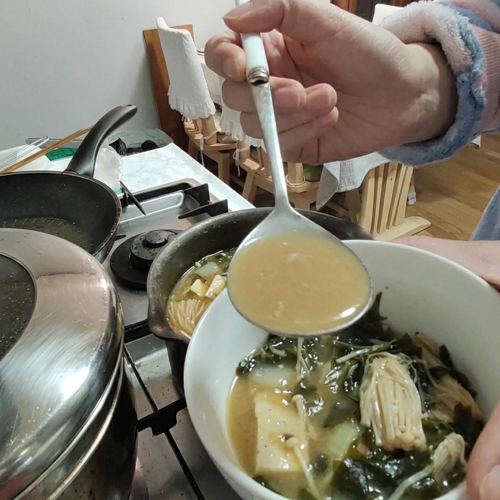 Pasta di soia aggiunta allo stufato con un mestolo, con tofu e funghi enoki visibili nella ciotola.