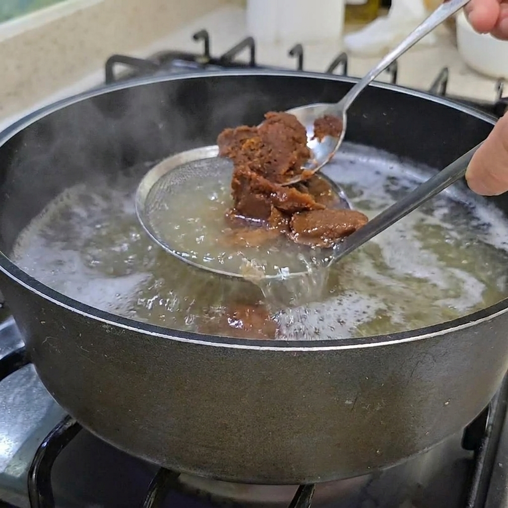 Due cucchiai di densa pasta di soia coreana marrone mescolati in una pentola di brodo chiaro.