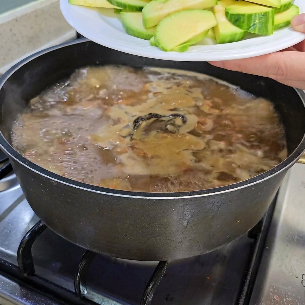 Zucchine affettate e un mazzetto di funghi enoki aggiunti alla pentola dello stufato.