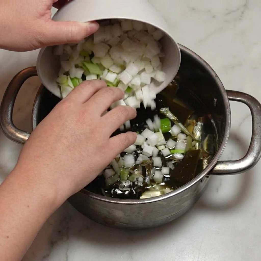 Daikon a cubetti e cipollotto tagliato versati da una ciotola in una pentola con acciughe e kombu.