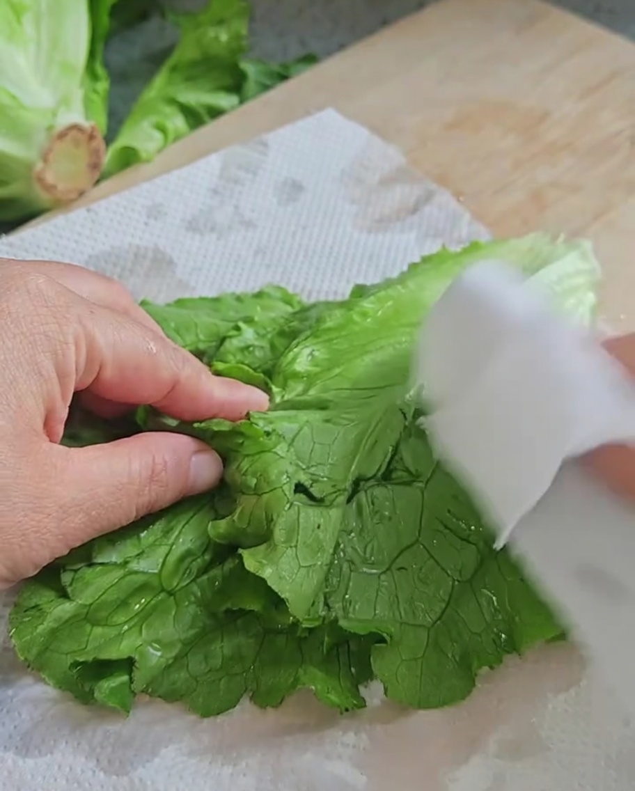Mani che asciugano foglie di lattuga fresca con carta assorbente su un tagliere in legno.