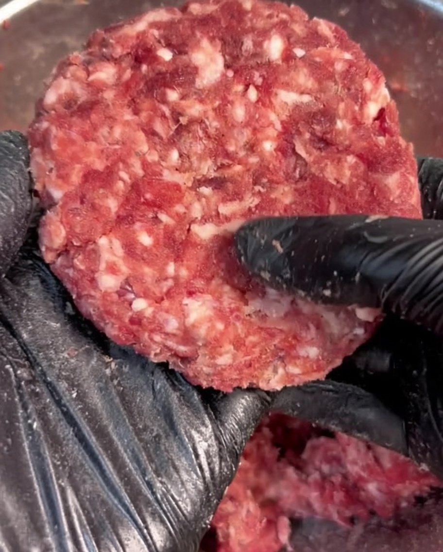 Mano con guanto nero che modella la carne macinata condita in una forma rotonda.