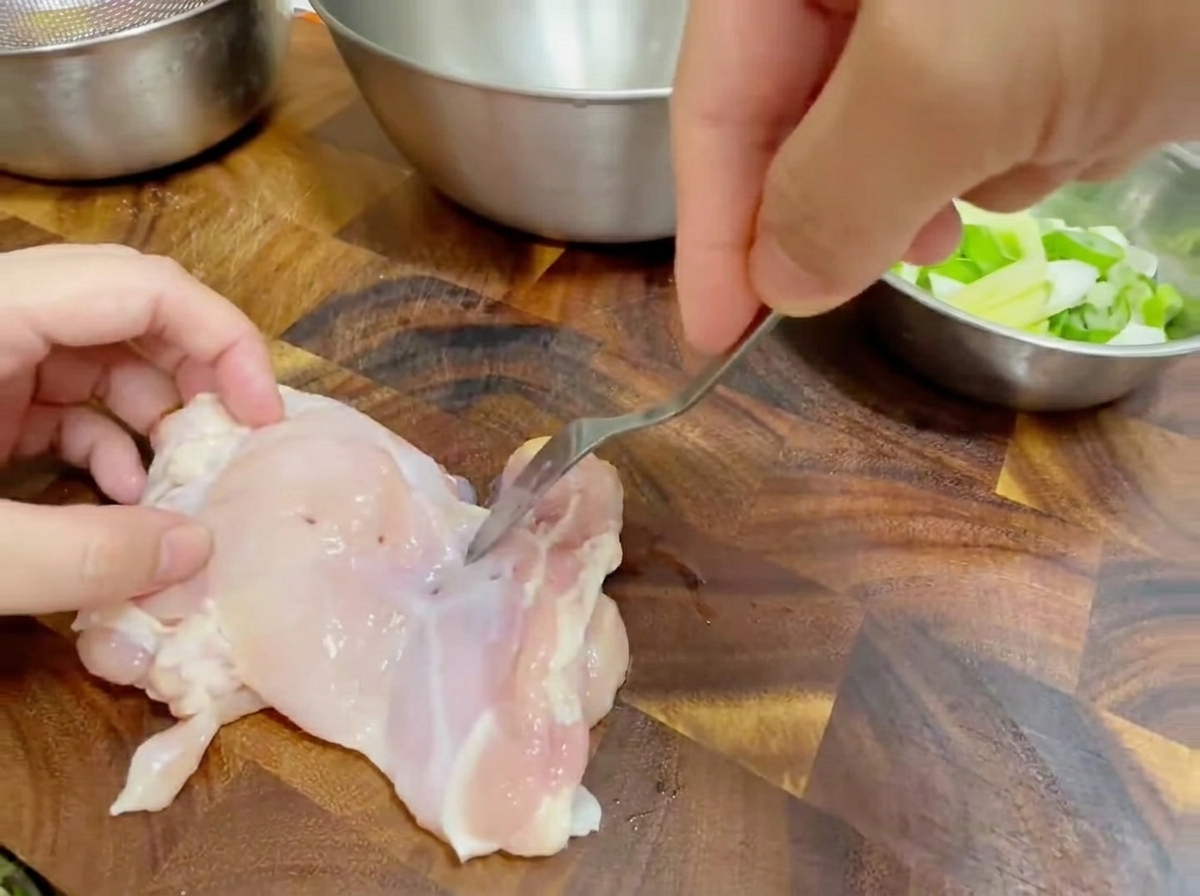 Primo piano di una forchetta che buca una sovracoscia di pollo cruda su un tagliere di legno.