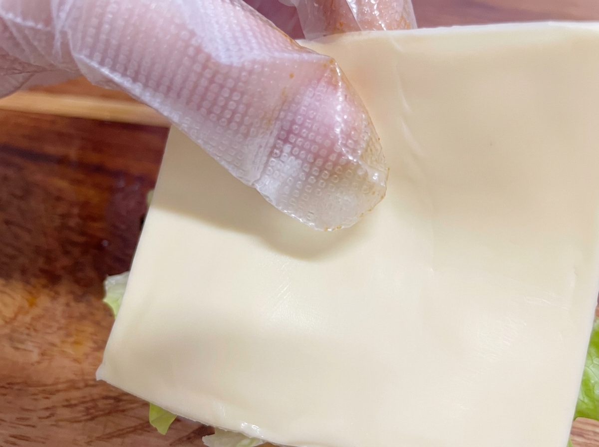 Una mano con guanto che posiziona una fetta di formaggio bianco sullhamburger assemblato.