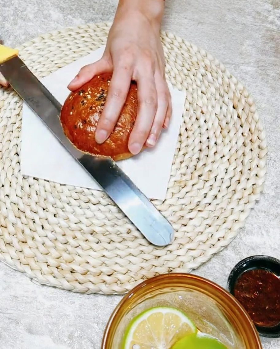 Una mano che utilizza un coltello per tagliare a metà un soffice pane per burger fatto in casa su un tagliere di legno.
