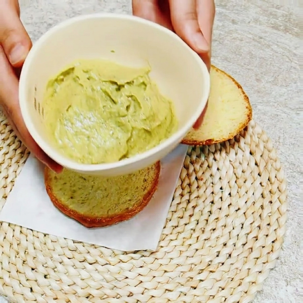Primo piano di un coltello che spalma la salsa verde brillante di avocado e basilico sullinterno del pane.