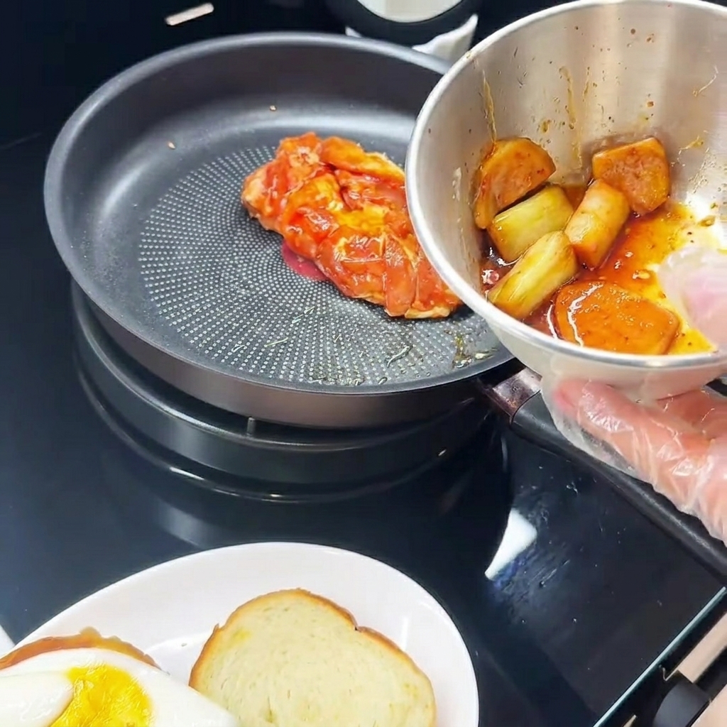 Coscia di pollo marinata in cottura in padella, lato pelle verso il basso, accanto a un piatto con un uovo fritto.