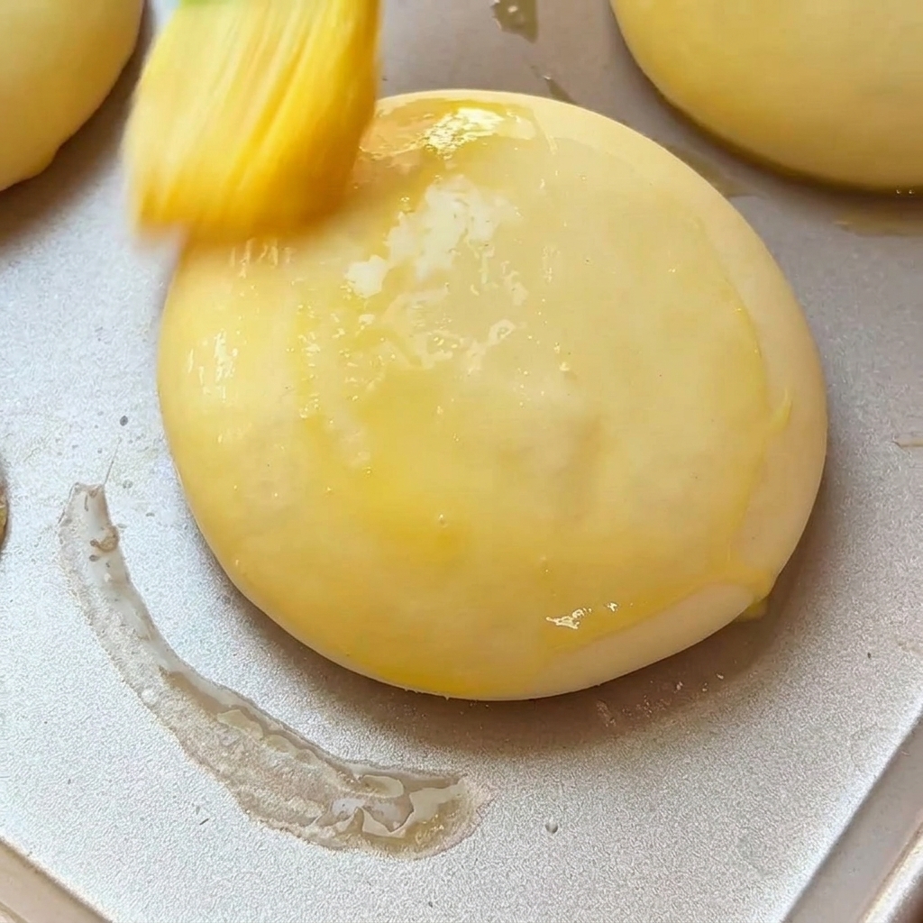 Un pennello da pasticceria che applica delluovo sbattuto su un panino da burger lievitato su una teglia di metallo.
