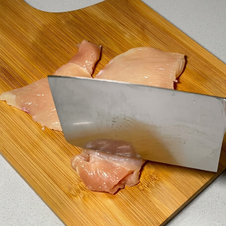 Un coltello da cucina utilizzato per battere le fette di pollo crudo su una superficie di legno.